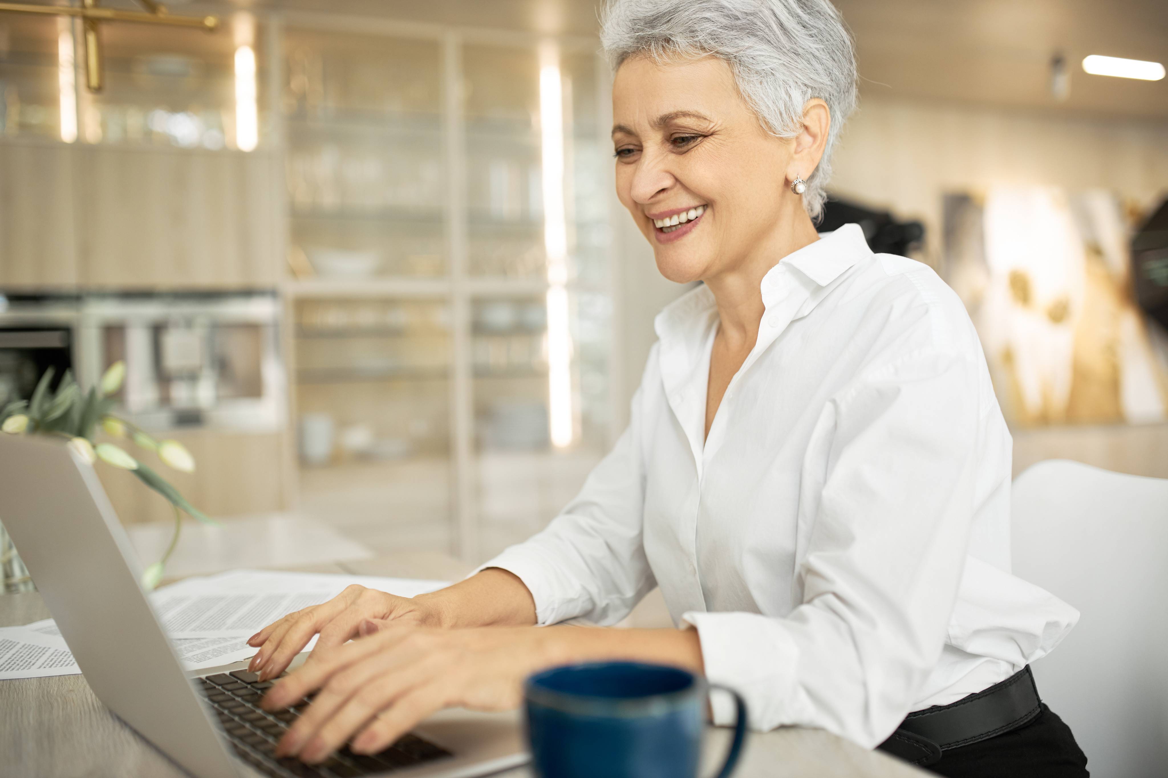 side view of happy middle aged businesswoman with short gray hair working on laptop in her stylish office with hands on keyboard typing letter sharing good news